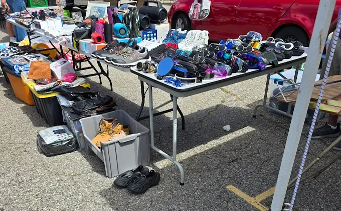 raynham flea market scene with tables displaying various items like shoes, sunglasses, electronics, and bags. a red car is visible in the background. bright, sunny day.