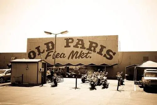 sepia-toned image of the old paris flea mkt building. motorcycles are parked in front, and a small booth and cars are visible nearby