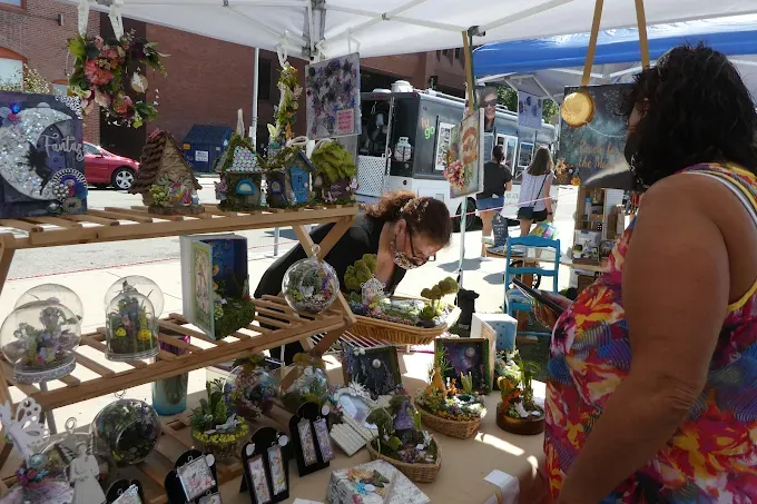 providence flea 2025 markets at craft fair stall under a canopy, showcasing handmade miniatures and decorative items. a woman browses while another arranges items.
