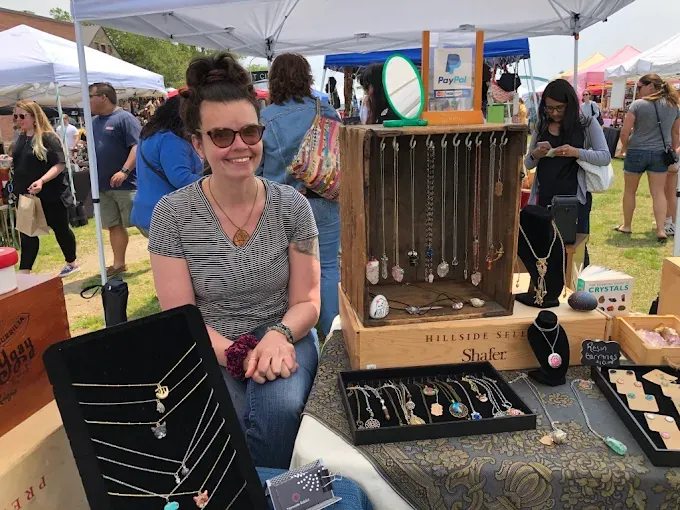providence flea 2025 markets at smiling vendor with sunglasses sits at a jewelry stall under a white tent at an outdoor market.