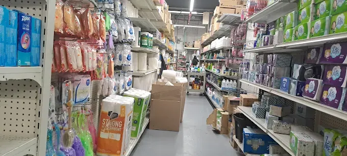 narrow store aisle with shelves stacked with household items like toilet paper, cleaning products, and paper towels. bright lighting and organized displays.