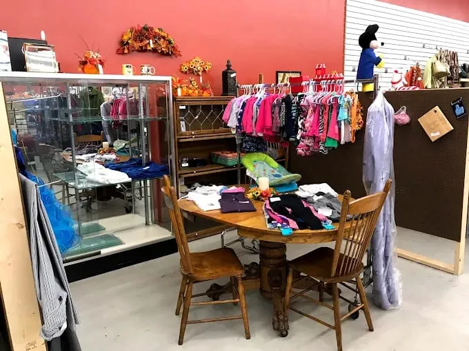 a funky munky flea market corner features a wooden table with chairs displaying folded clothes. a clothing rack with colorful children's outfits and autumn decor adorns the wall.