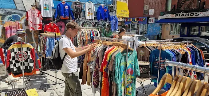 a man browses colorful vintage clothing on outdoor racks in a urban street market. a graffiti wall and shop signs create an eclectic, vibrant atmosphere at ludlow flea market