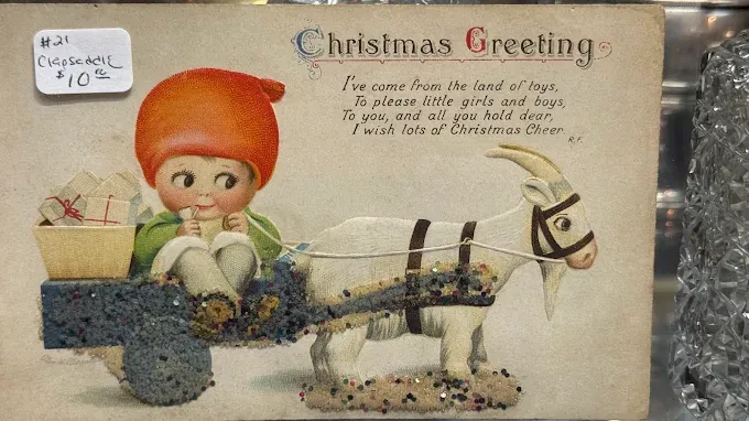 a vintage christmas greeting postcard featuring a child in a red hat riding a cart pulled by a goat.
