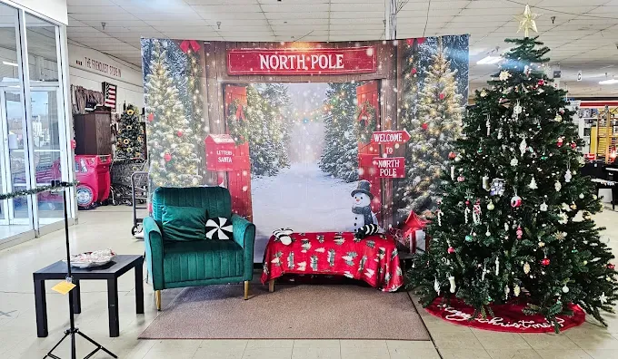 festive christmas setup with a north pole backdrop, green chair, red and white blanket on a bench, decorated christmas tree, and holiday signs, evoking a cheerful holiday spiritat dover flea market