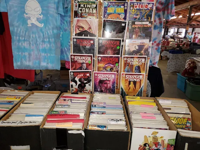 french 500 flea market rows of comic books are displayed in boxes, with stranger things editions prominently featured. a blue tie-dye shirt hangs above, creating a vibrant, nostalgic scene.
