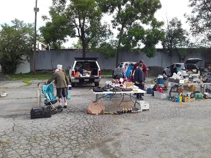 outdoor flea market with tables of assorted items. people browse under scattered trees in a cracked parking lot, creating a casual, laid-back atmosphere.