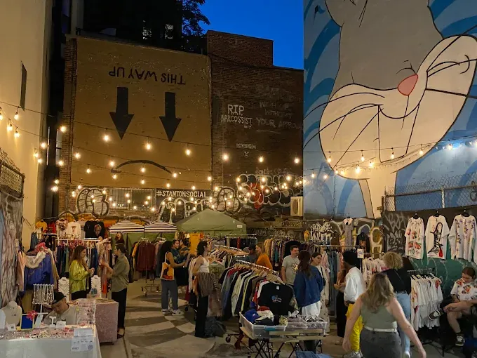outdoor night market with string lights, vibrant street art, and people browsing clothing racks. a mural of a cartoon character adds a playful atmosphere at ludlow flea market